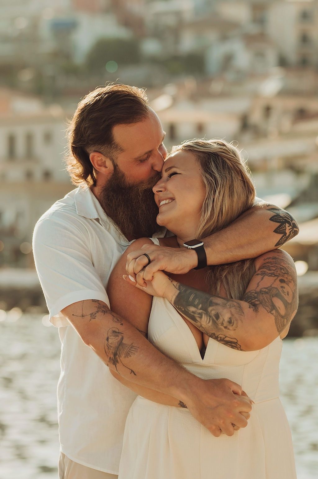 A bearded man with tattoos kisses a smiling tattooed blonde woman in white dress, embracing outdoors near a sunny coastal town.