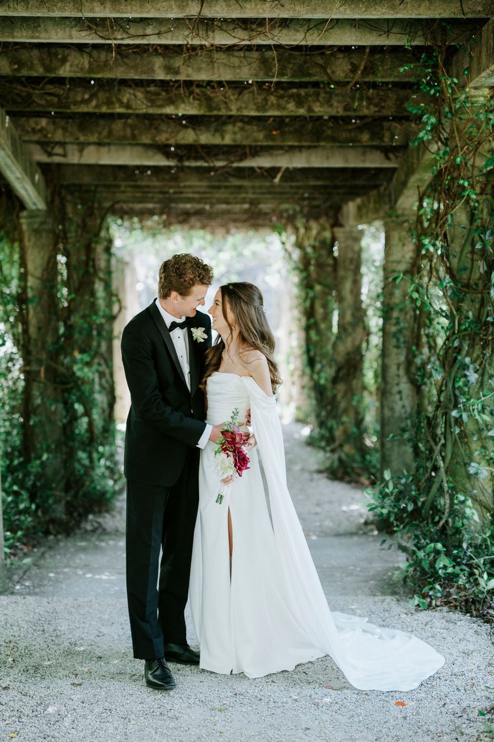 Bride and groom share romantic moment under ivy-covered pergola, bride in off-shoulder gown holding burgundy bouquet.