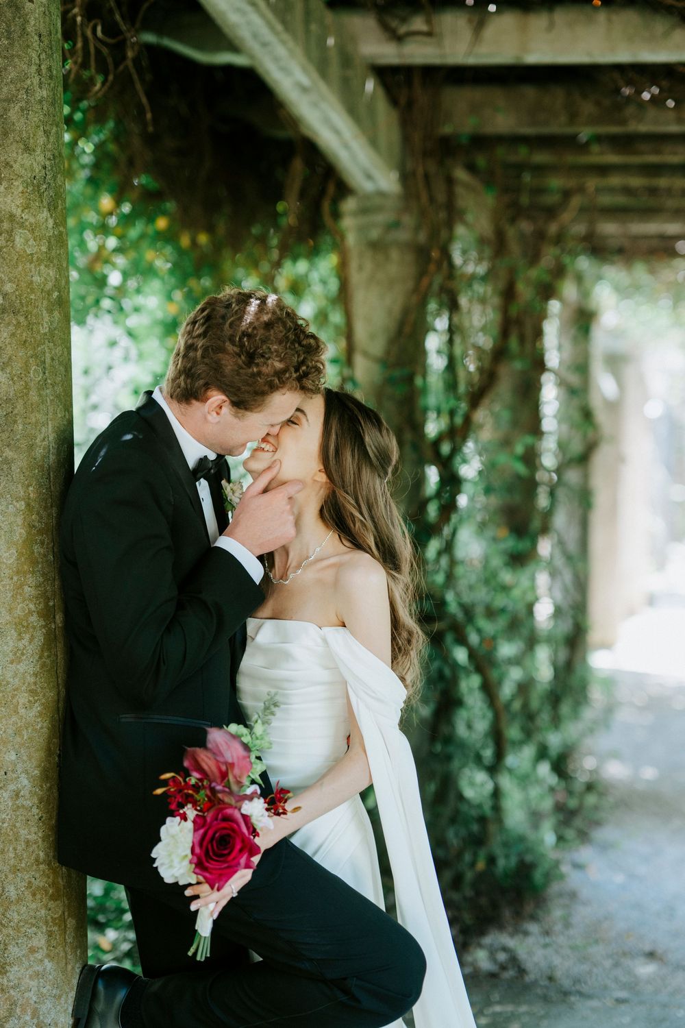 Bride and groom share a romantic moment under a vine-covered pergola, bride holding a red and white bouquet.