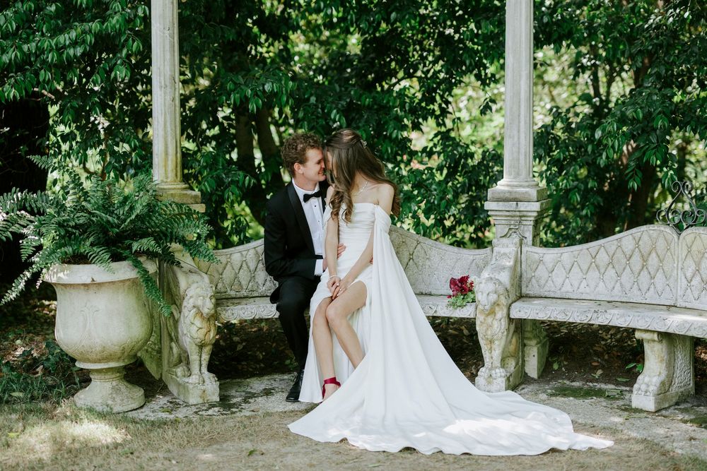 Bride and groom share a romantic moment on ornate stone bench in lush garden with classical columns.