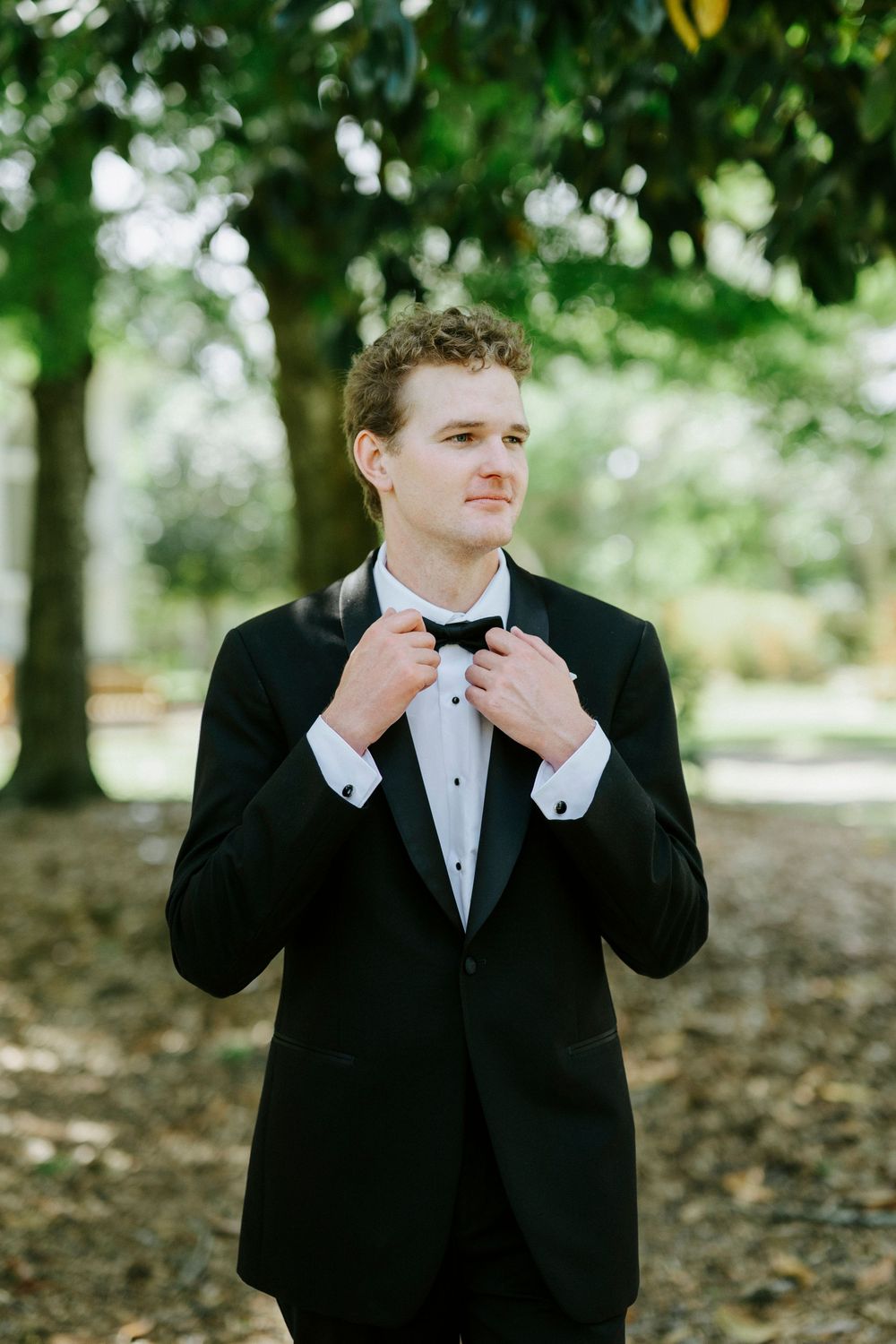 A groom in a black tuxedo adjusts his bow tie outdoors under lush green trees on his wedding day.