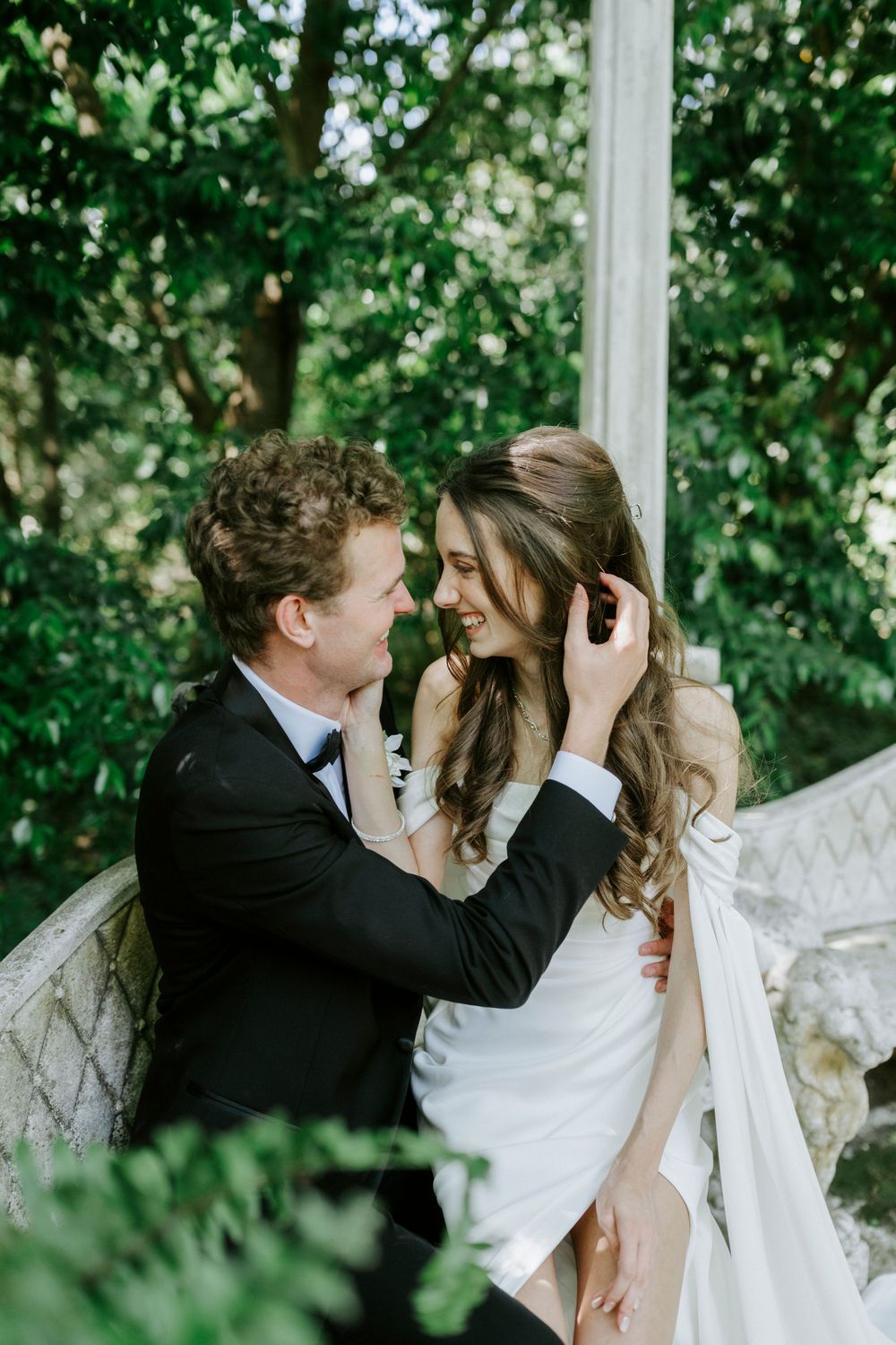 A groom in a black tuxedo tenderly touches his smiling bride's hair outdoors amid lush green trees.