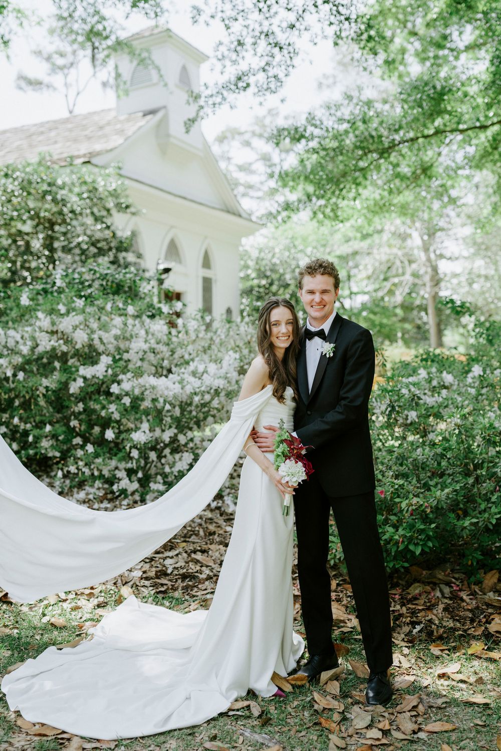 Bride and groom pose outside a white chapel surrounded by lush greenery, her veil flowing dramatically in the breeze.