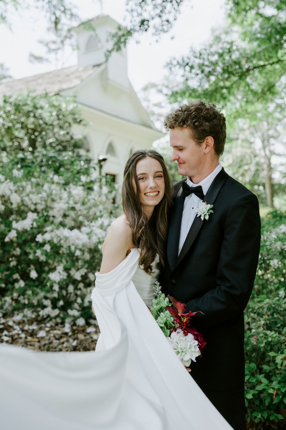 Bride in off-shoulder gown and groom in black tuxedo smile together outdoors near a white chapel surrounded by blooming trees.