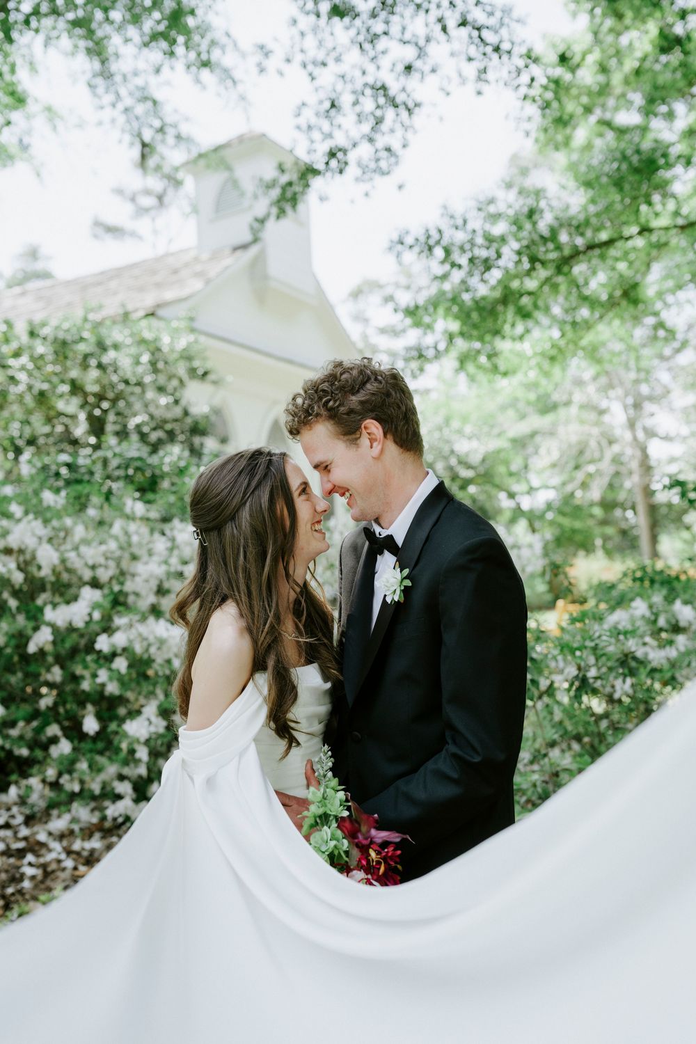 Bride and groom share a romantic moment outdoors, surrounded by lush greenery, with flowing veil and red bouquet.