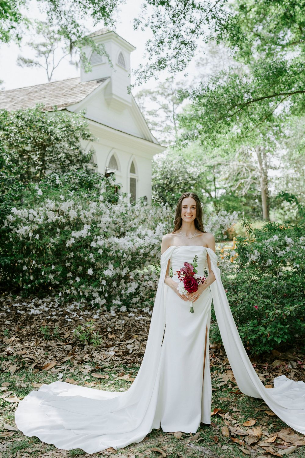 Bride in off-shoulder white gown with cape holds red bouquet before a white chapel surrounded by blooming greenery.