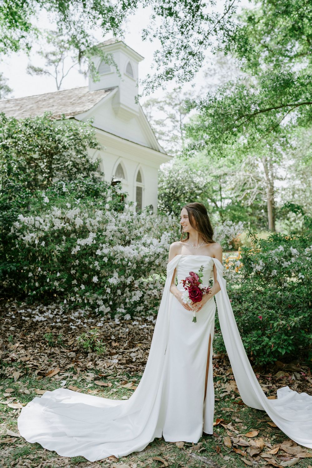Bride in off-shoulder white gown with cape sleeves holds bouquet before a white chapel surrounded by lush greenery.