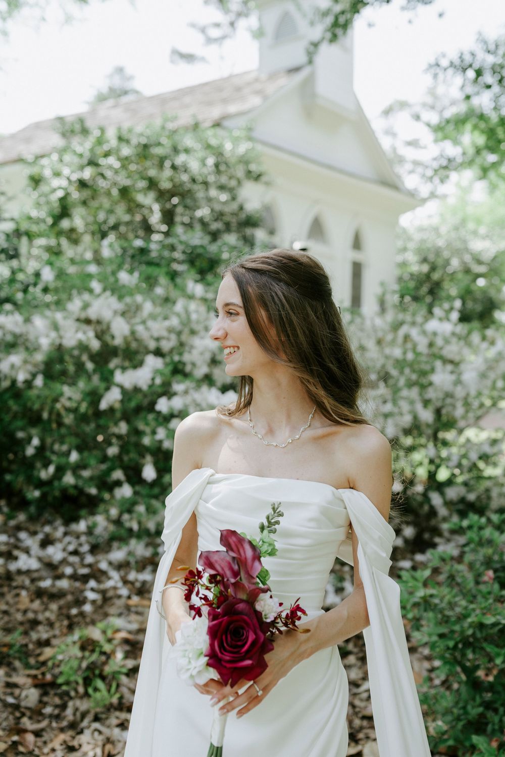 Smiling bride in off-shoulder white gown holds red rose bouquet outdoors near white chapel surrounded by flowering bushes.