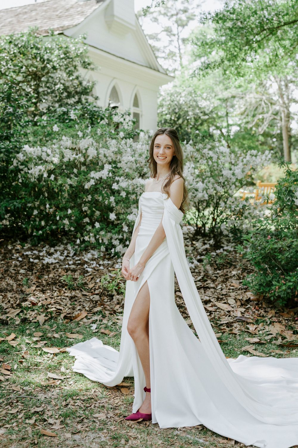 Smiling bride in white off-shoulder gown with slit and train poses outdoors near a white chapel surrounded by blooming trees.