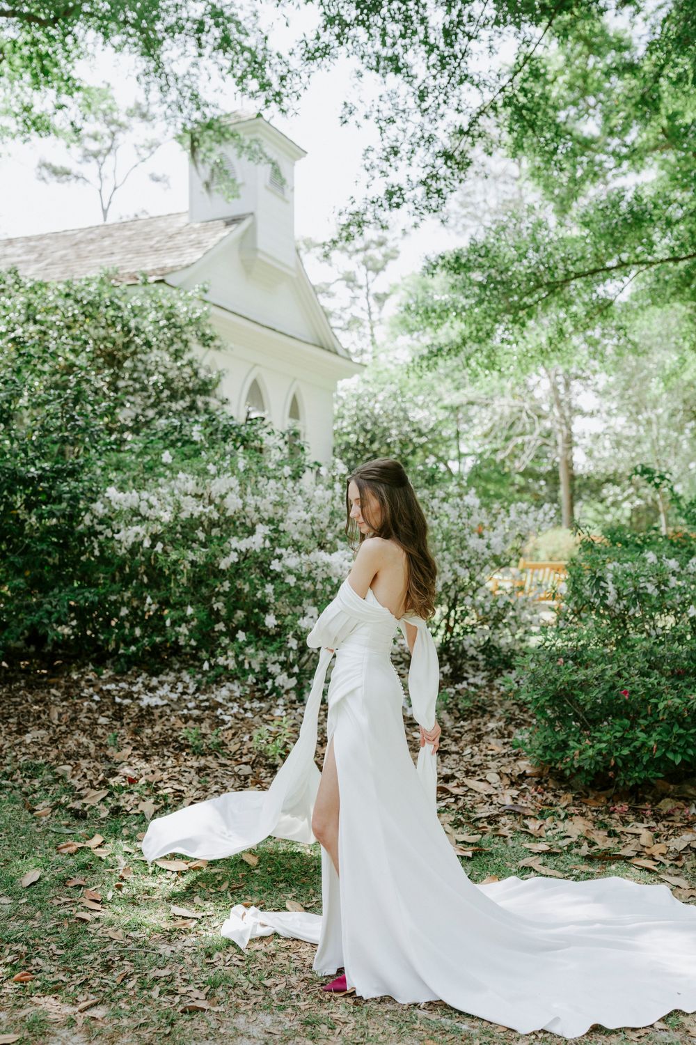 Elegant bride in a white off-shoulder gown with long train poses outdoors near a white chapel amid green trees.