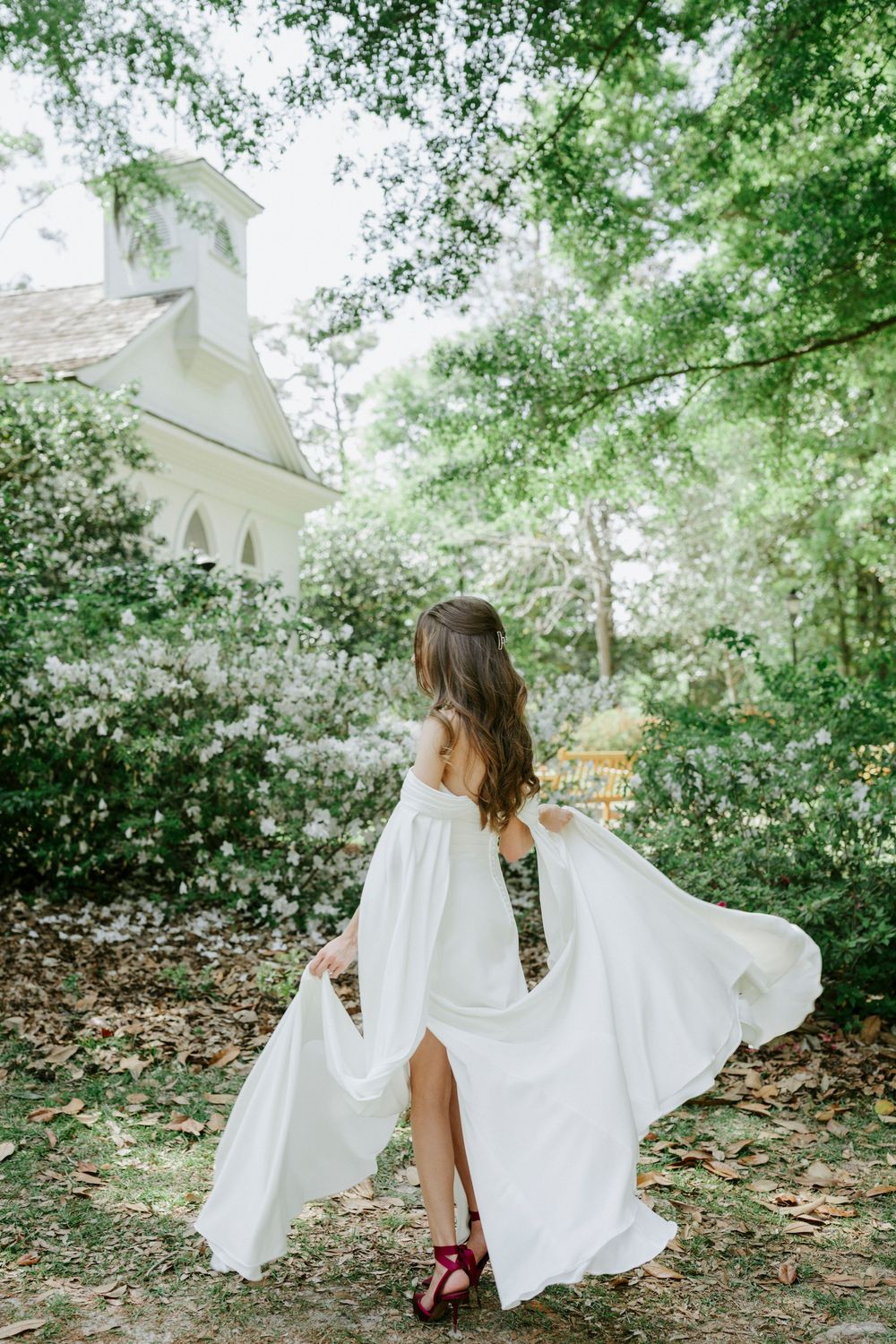 Bride in flowing white off-shoulder gown with dramatic sleeves twirling in lush garden near white chapel.