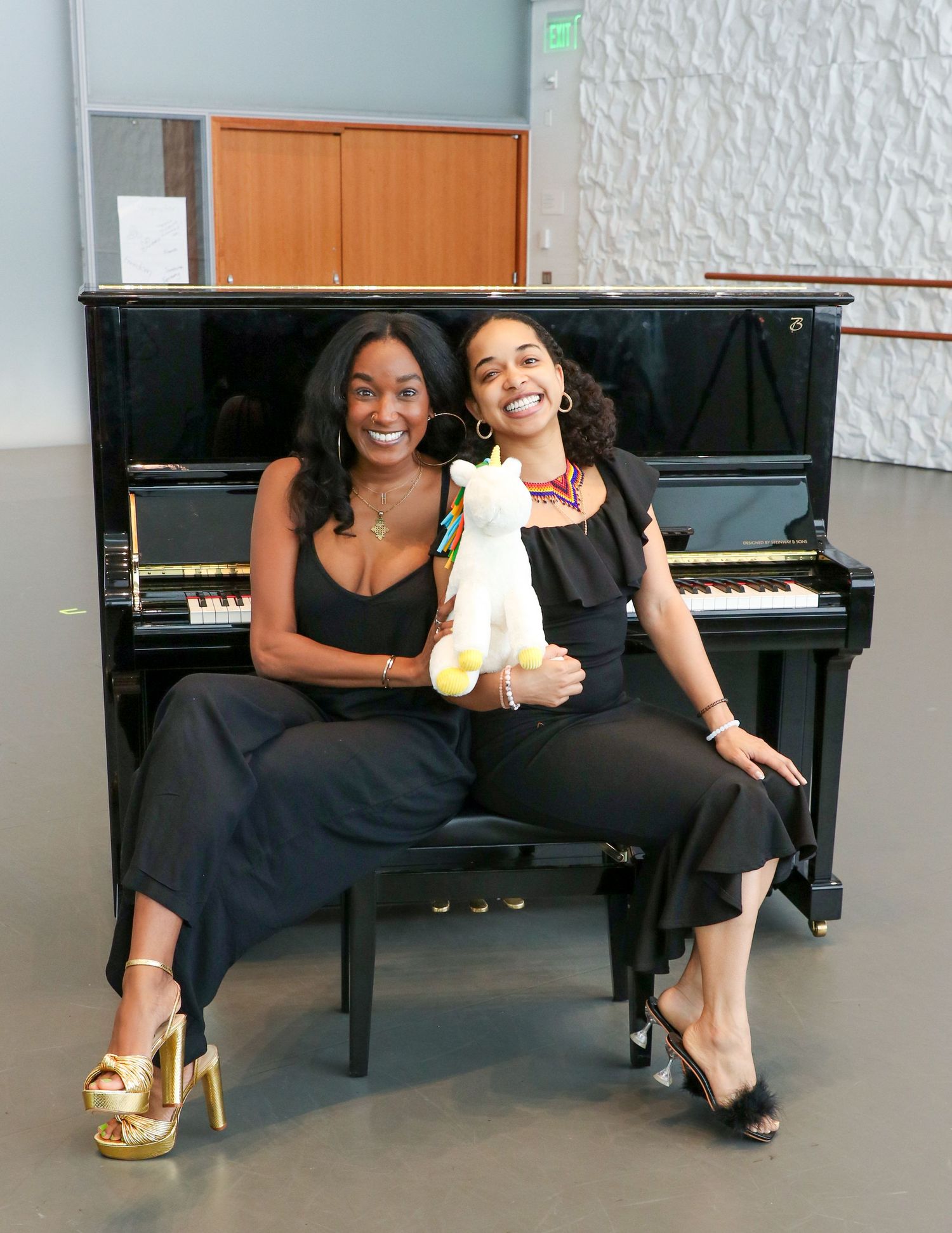 Five women in black outfits pose together around a piano in a modern interior space.