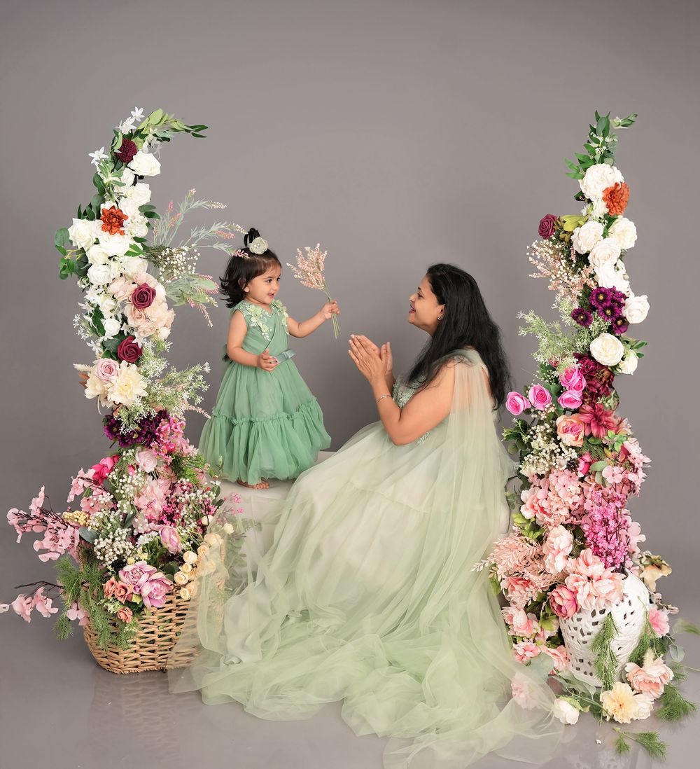 A mother and daughter in matching sage green dresses pose by floral arch with pink and white blooms in a studio.