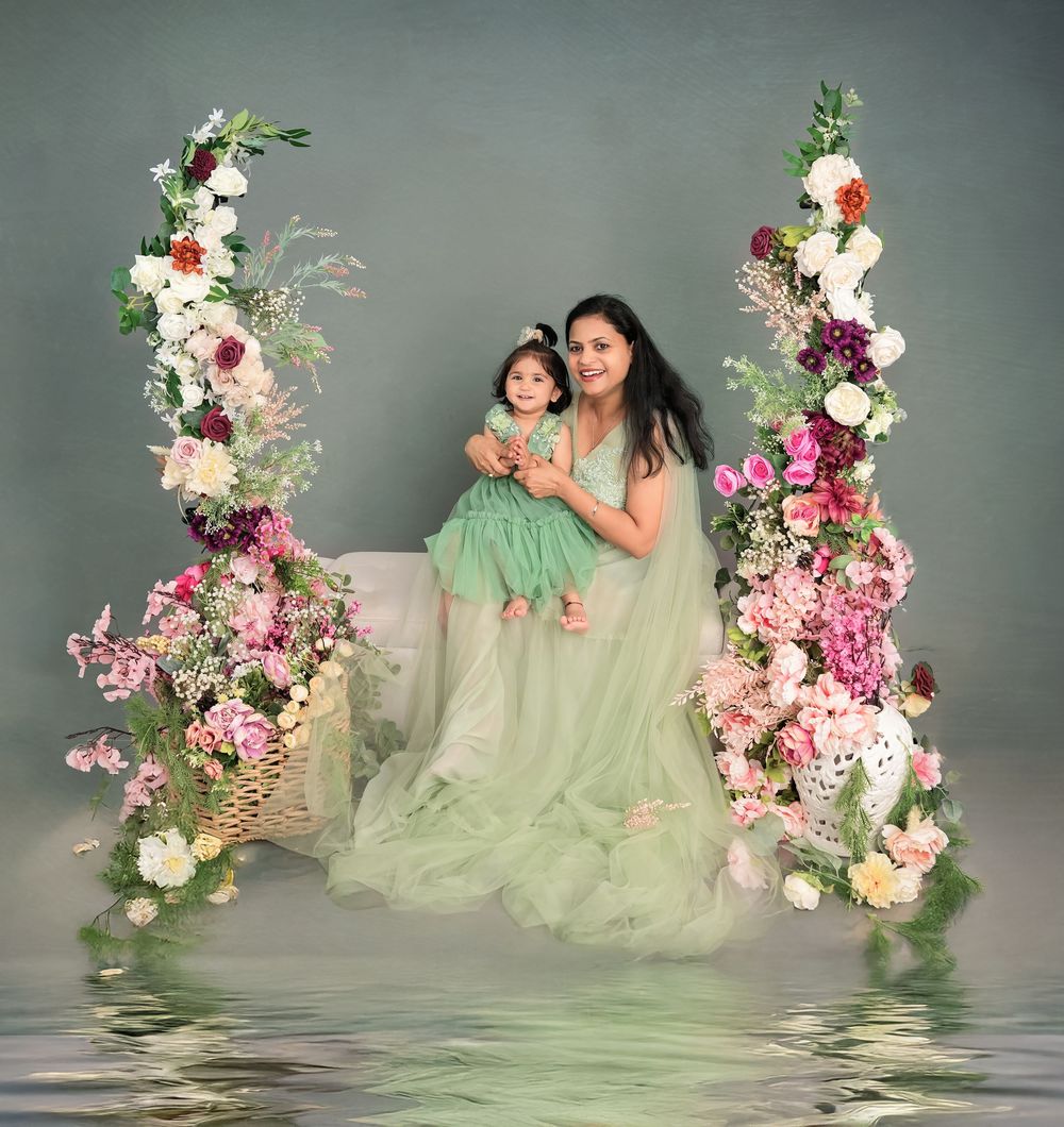 Mother and daughter in matching green dresses pose on a floral swing surrounded by pink and white blooms.