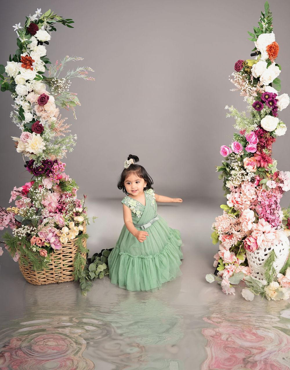 A young girl in a mint green dress poses among lush floral arrangements in a professional studio photo.