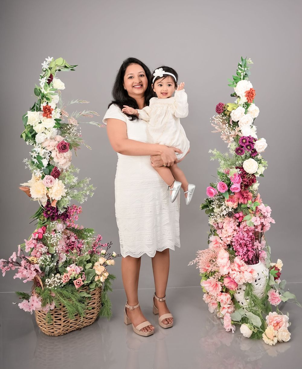Woman in white dress holds baby girl surrounded by floral arrangements in a studio portrait.