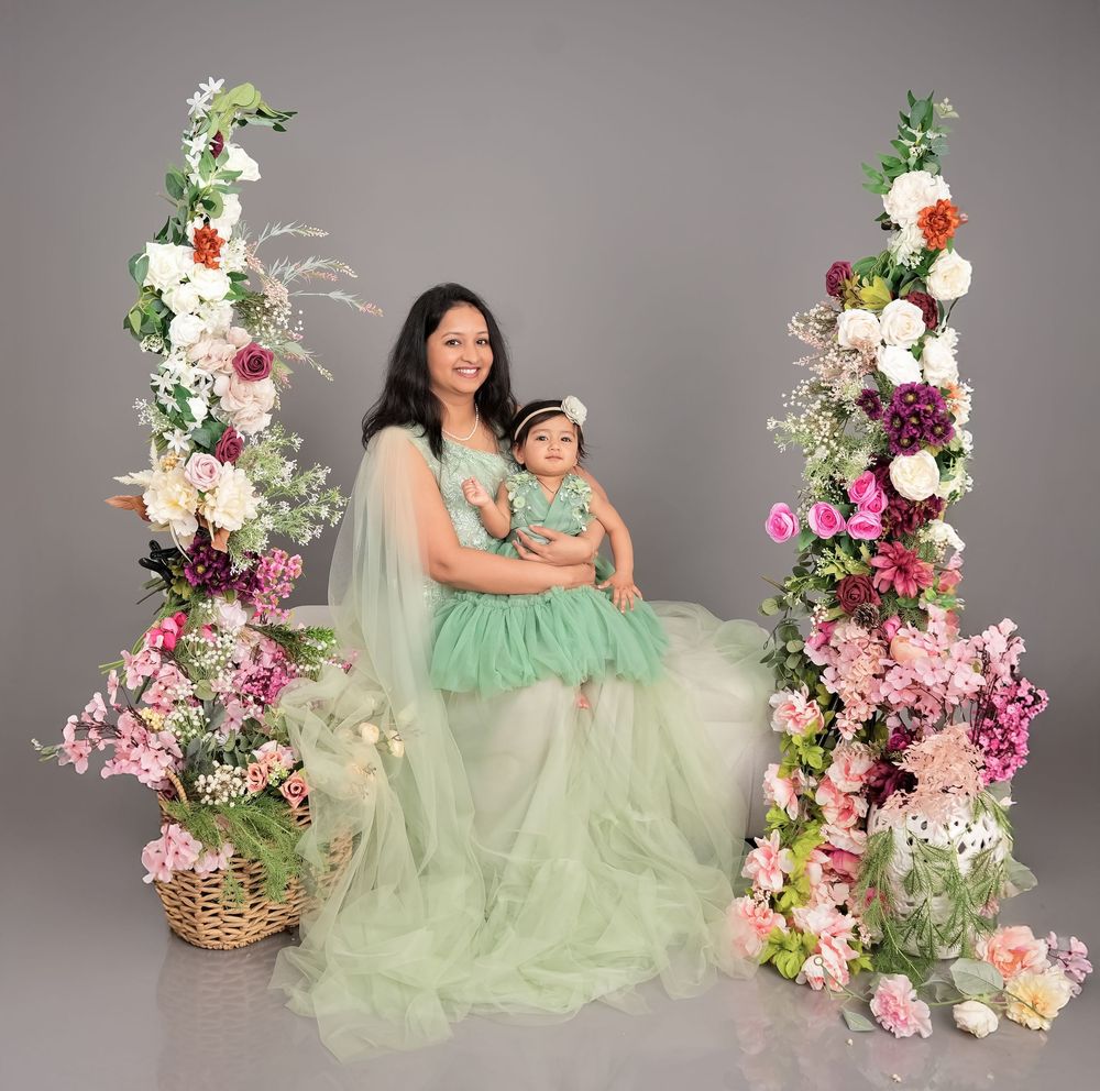 Mother and baby daughter in matching green tulle dresses pose among floral arrangements in a studio portrait.