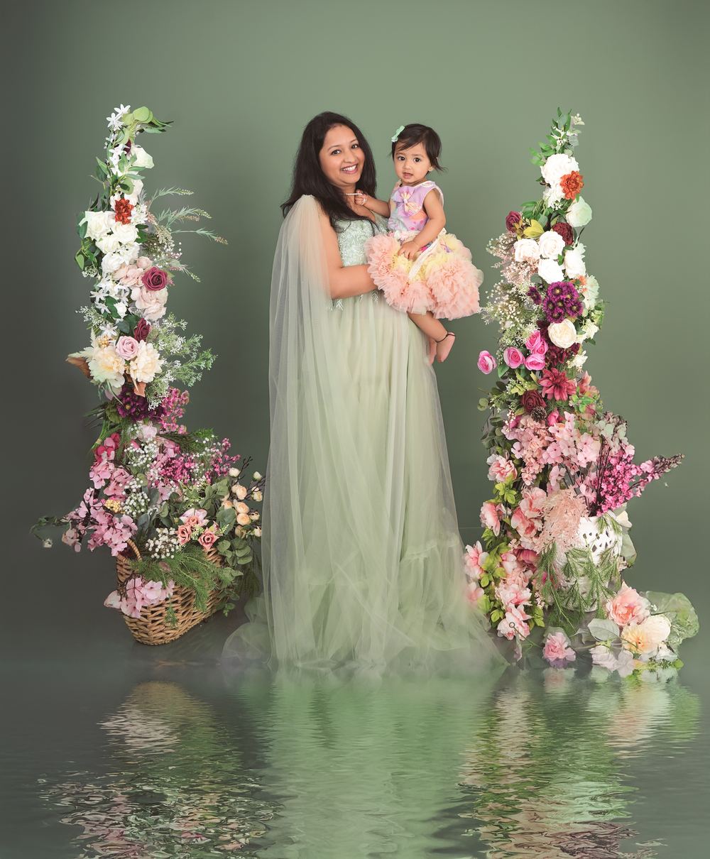 Mother in sage tulle gown holds toddler in pink dress between floral columns with pink and white blooms.