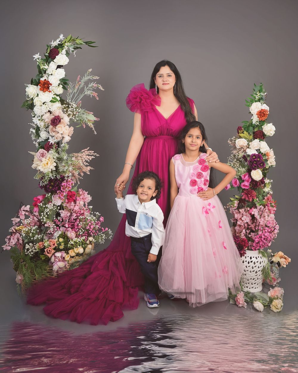 Woman in magenta gown poses with two children amid lush floral arrangements in a studio portrait.