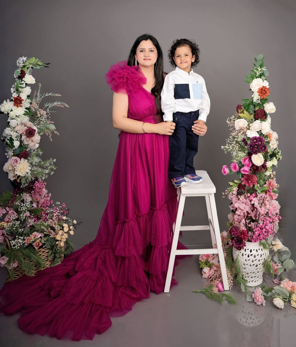 Woman in magenta gown poses with young boy in white shirt on white stool, surrounded by floral arrangements.