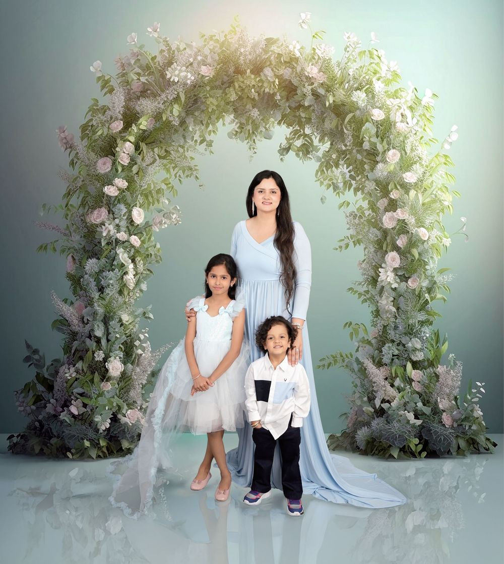 A woman in a light blue gown poses with two children under a floral arch with white roses and greenery.