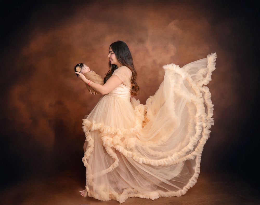 Woman in flowing cream ruffled gown holding camera, posing gracefully against warm brown studio backdrop.