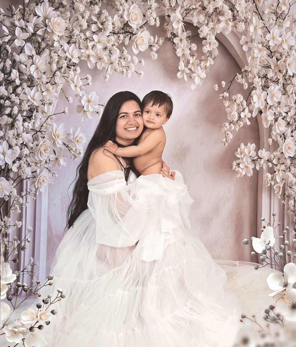 Woman in white gown holds smiling baby under floral arch with white roses and blossoms in elegant portrait.
