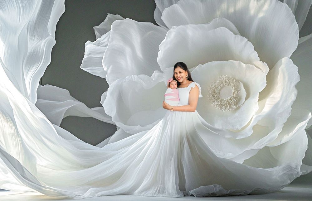 A mother in a white gown holds her baby amid a giant white floral backdrop in an elegant maternity photo.
