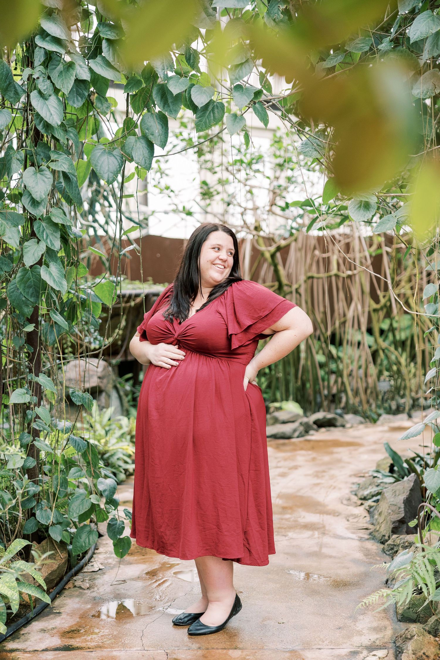 Radiant pregnant woman in burgundy dress poses with hands on hips beneath a lush green vine archway in a greenhouse.