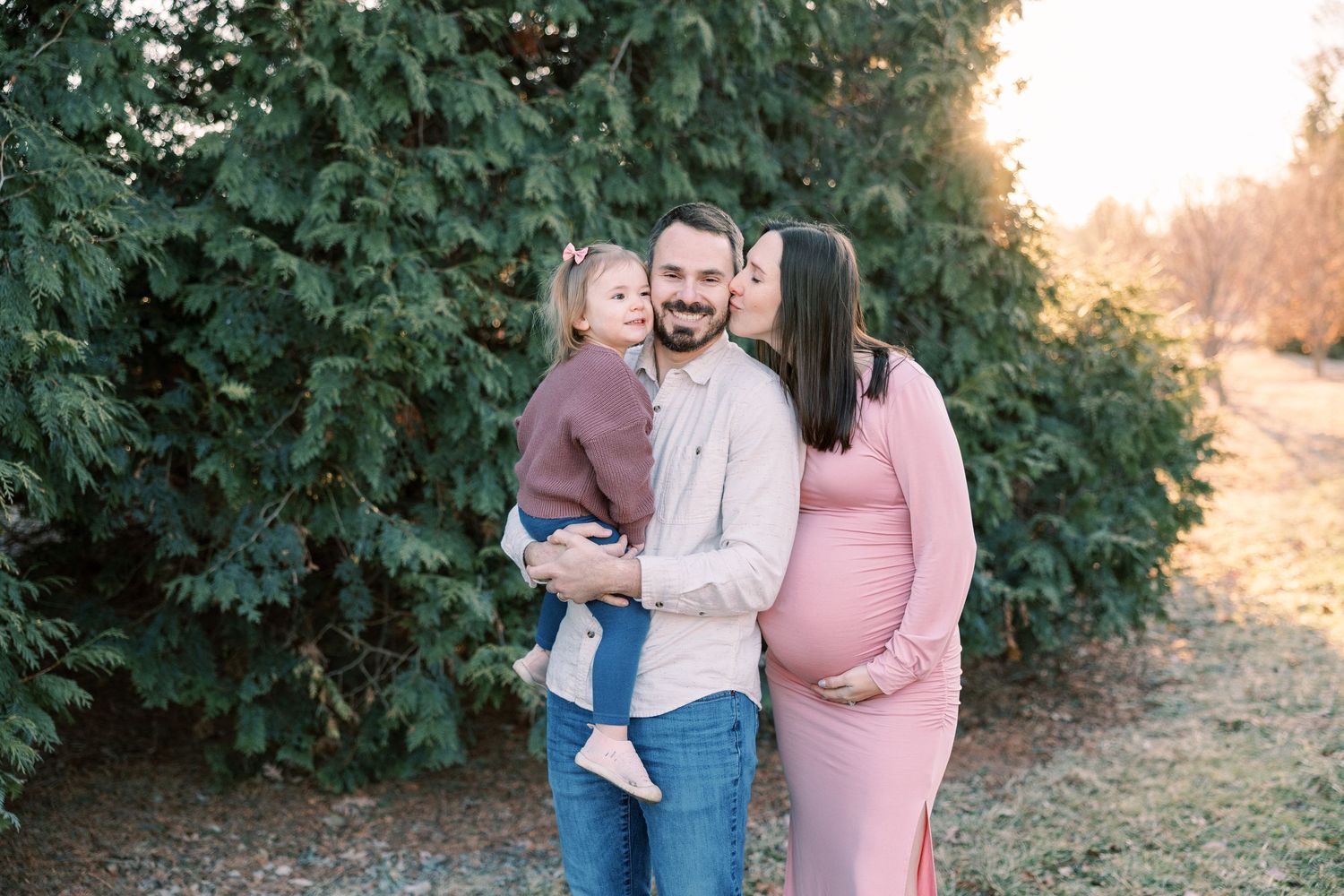 Happy family with toddler and pregnant mother sharing a kiss outdoors near evergreen trees.