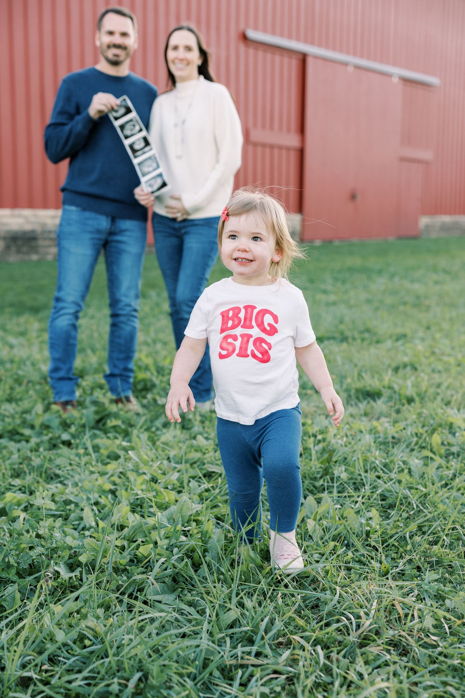 A toddler in a Big Sis shirt runs toward the camera while her parents hold ultrasound photos behind her.
