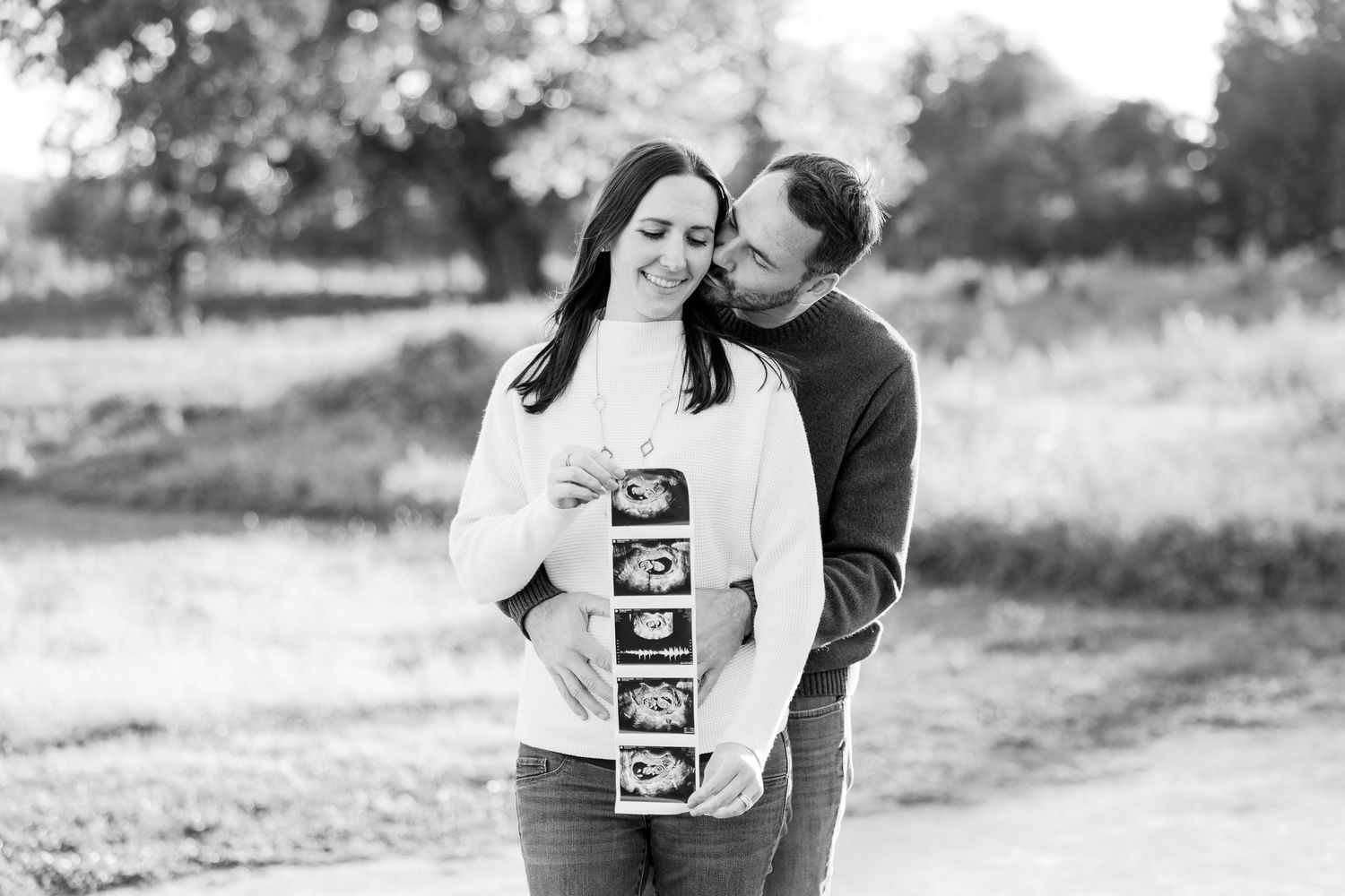 Black and white photo of couple holding ultrasound strip, smiling tenderly in a park setting.