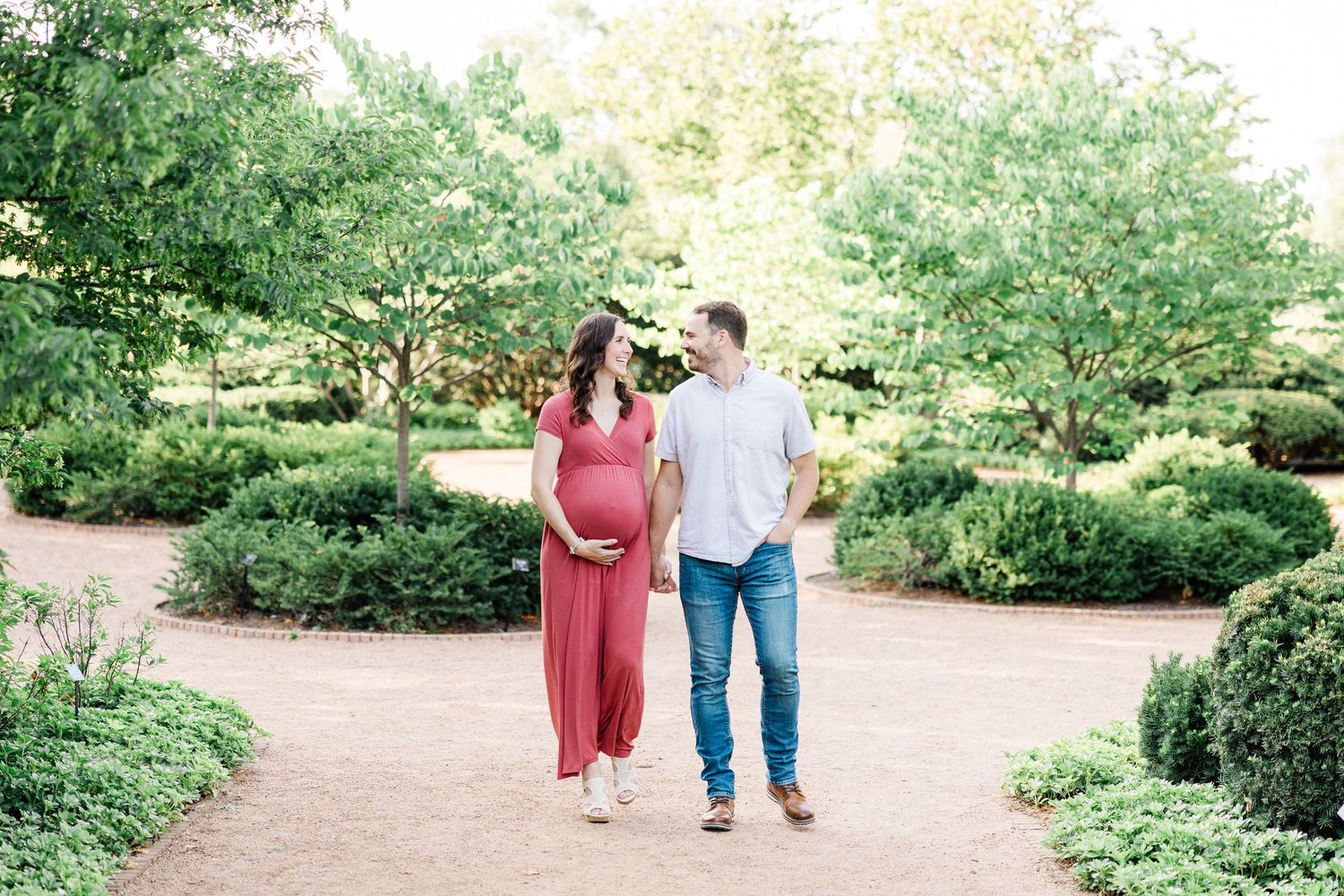 Pregnant woman in red maxi dress and partner walk along garden path lined with green trees.