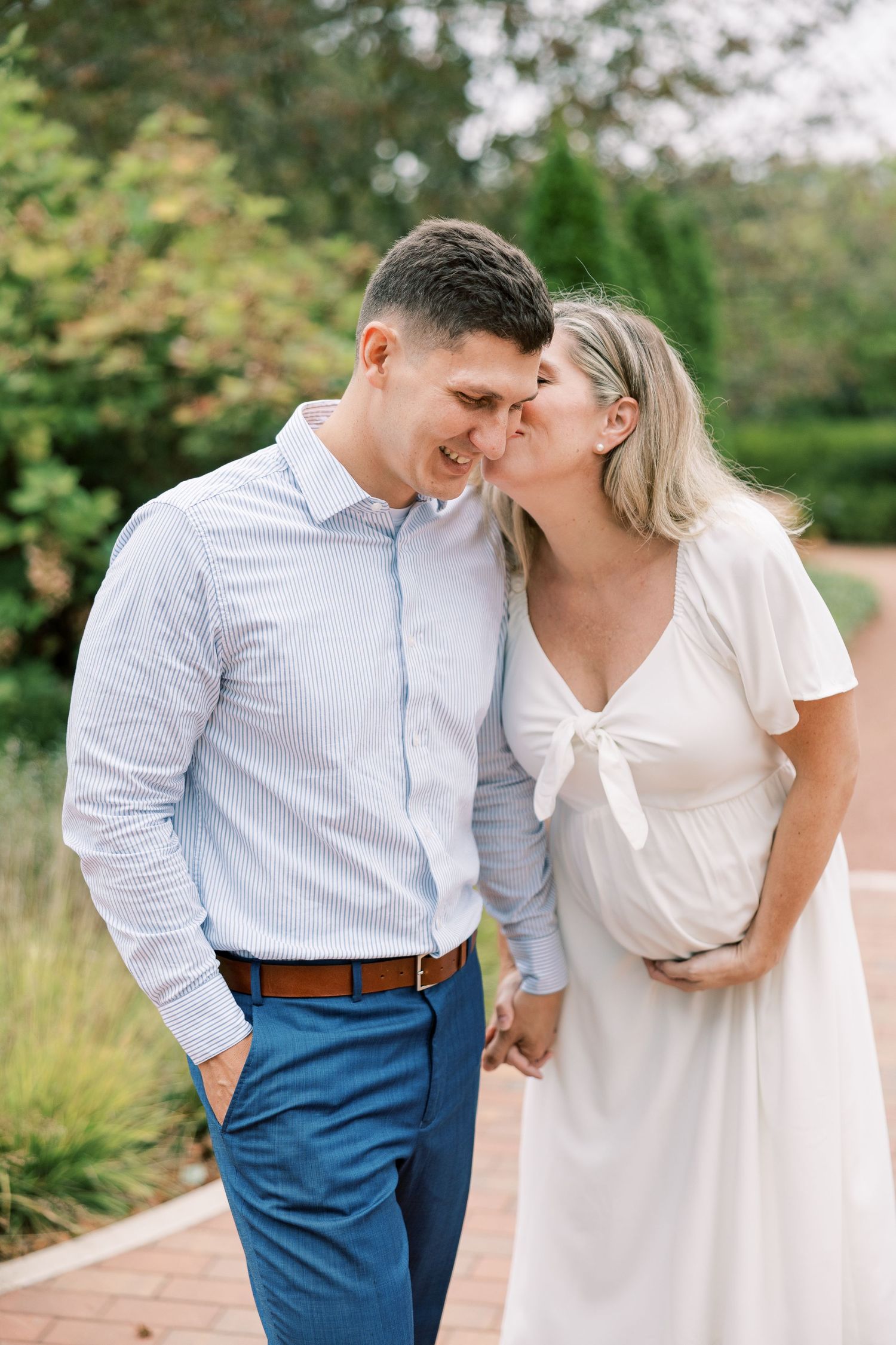 Pregnant woman in white dress kisses man in blue shirt and pants outdoors in garden setting.