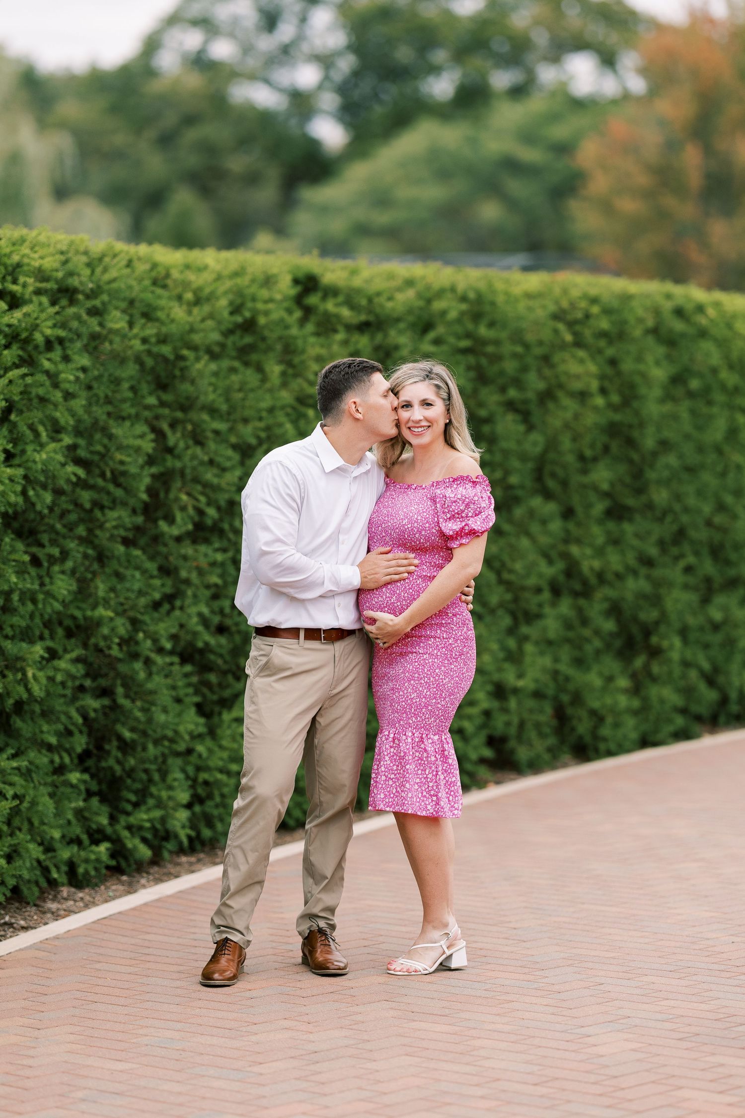 A couple poses on a brick path, man kissing pregnant woman in pink floral dress near green hedges.