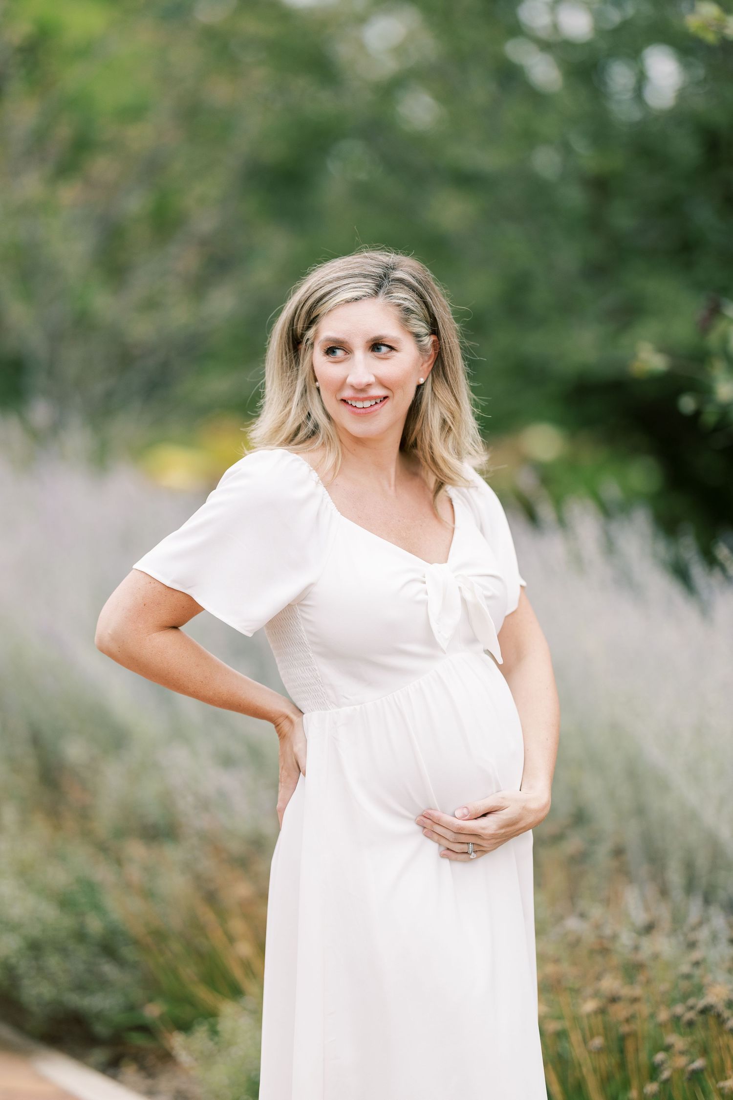 Smiling pregnant woman in white flutter-sleeve dress cradles baby bump in blooming garden.