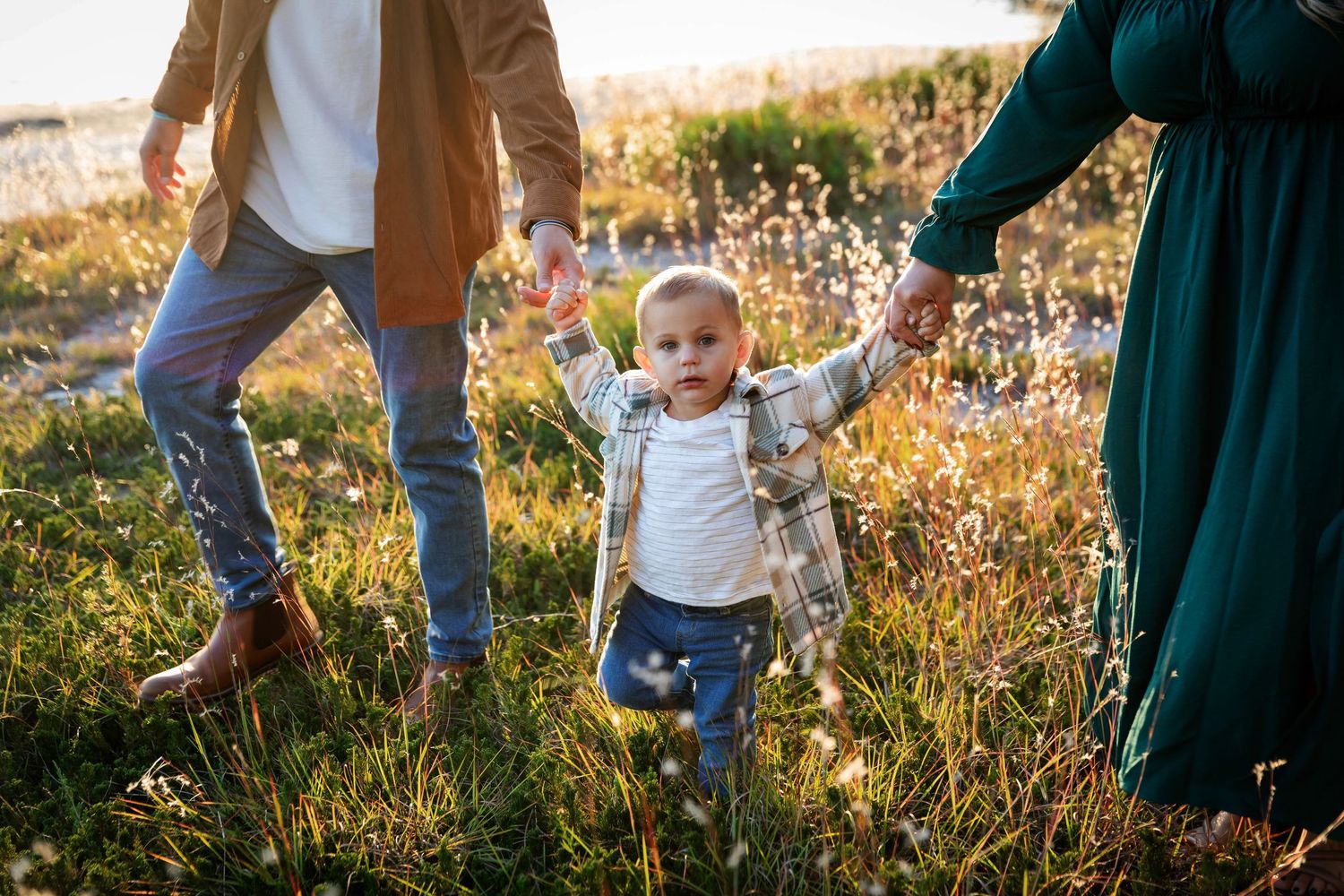 Young toddler walks hand-in-hand with parents through a sunny wildflower meadow in a heartwarming family photo.