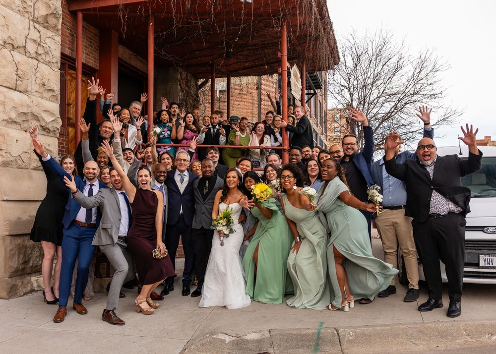 Joyful wedding party and guests celebrating outside a rustic stone building, bride in white holding yellow flowers.
