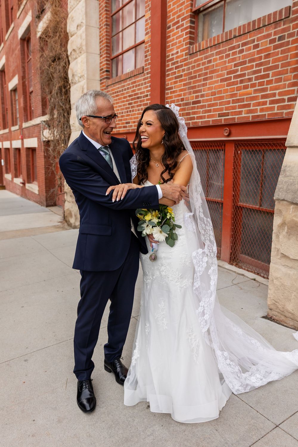 Bride in white lace gown laughing with older man in navy suit, holding yellow bouquet outside brick building.