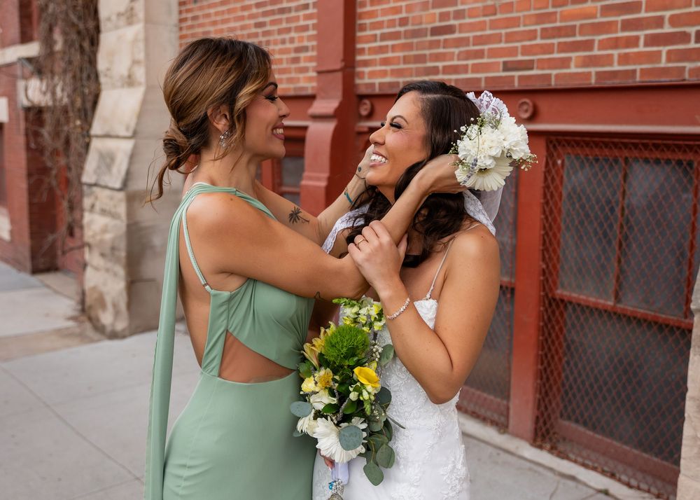 Bridesmaid in sage green dress adjusts brides hair while holding bouquets outside brick building.