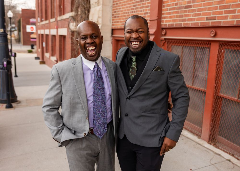 Two well-dressed men in suits laughing together on a city sidewalk with brick buildings in the background.