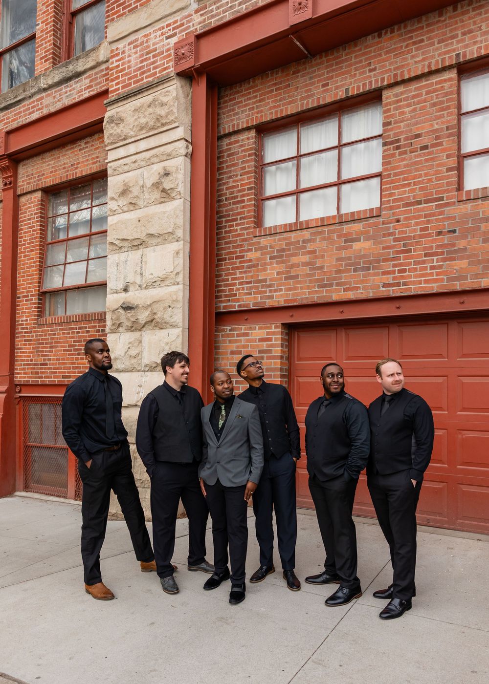 Six men in dark formal attire pose together outside a red brick building.