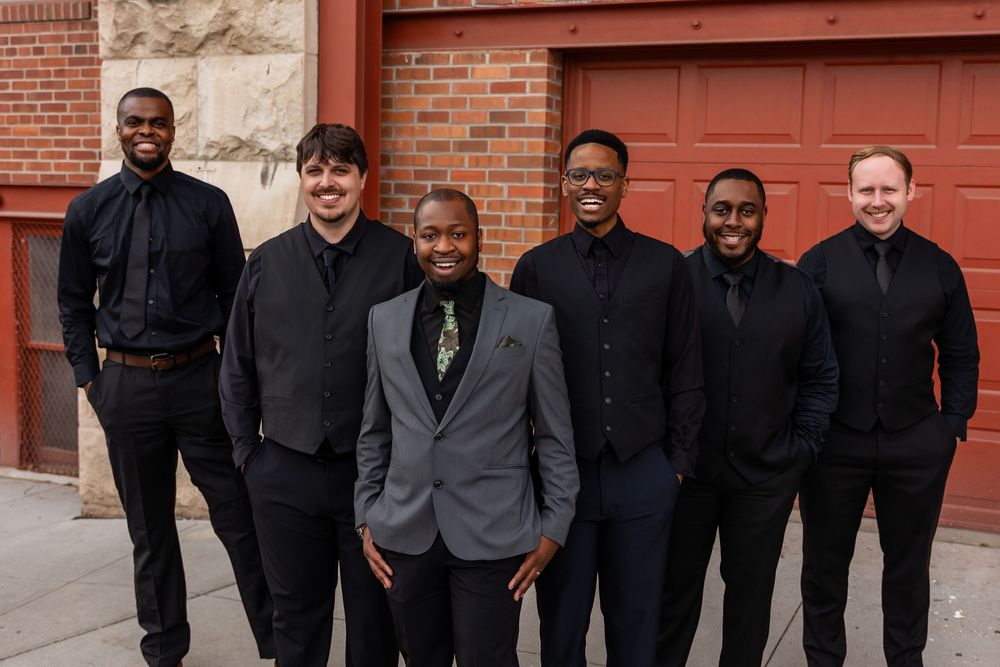 Six young men in formal black attire smile together outdoors near a brick building.