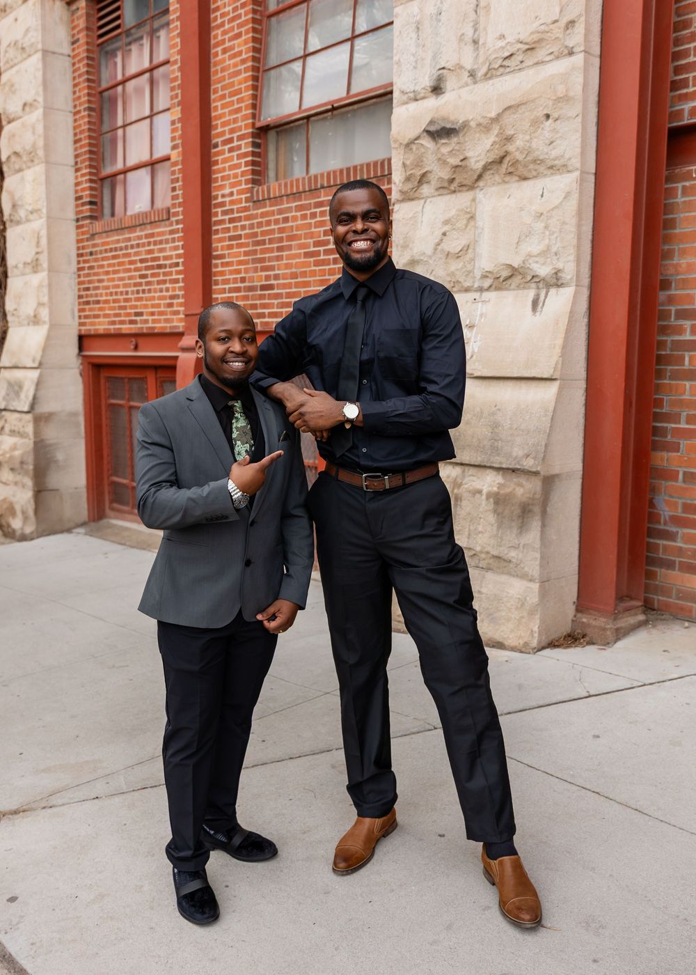Two well-dressed men in suits smile and pose together outside a historic brick building.