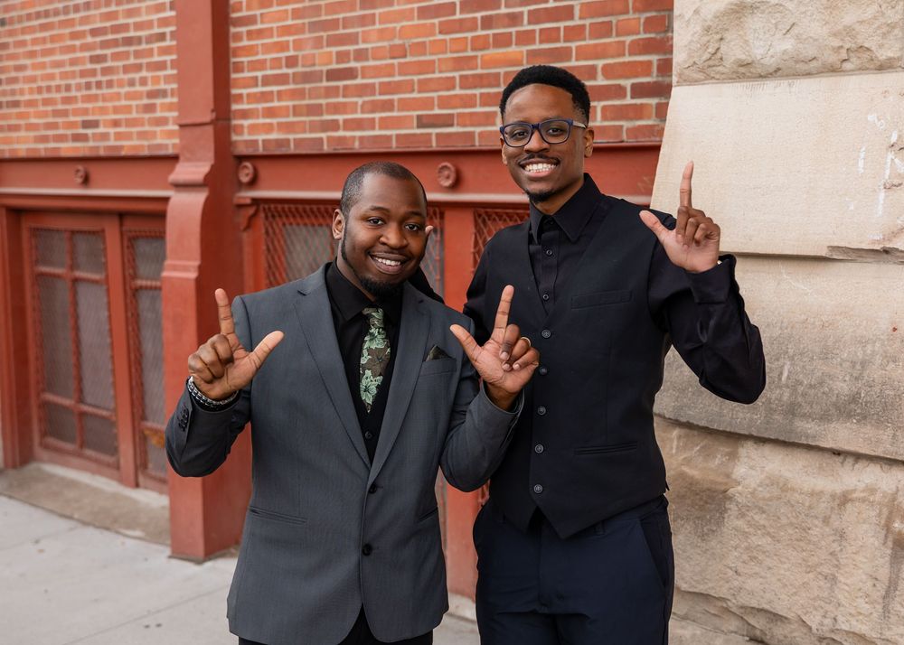 Two well-dressed men smiling and posing with hand gestures in front of a brick building.