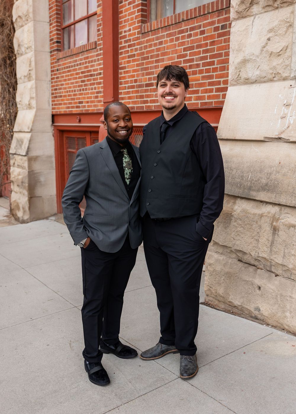 Two men in formal attire smile together outside a red brick building.