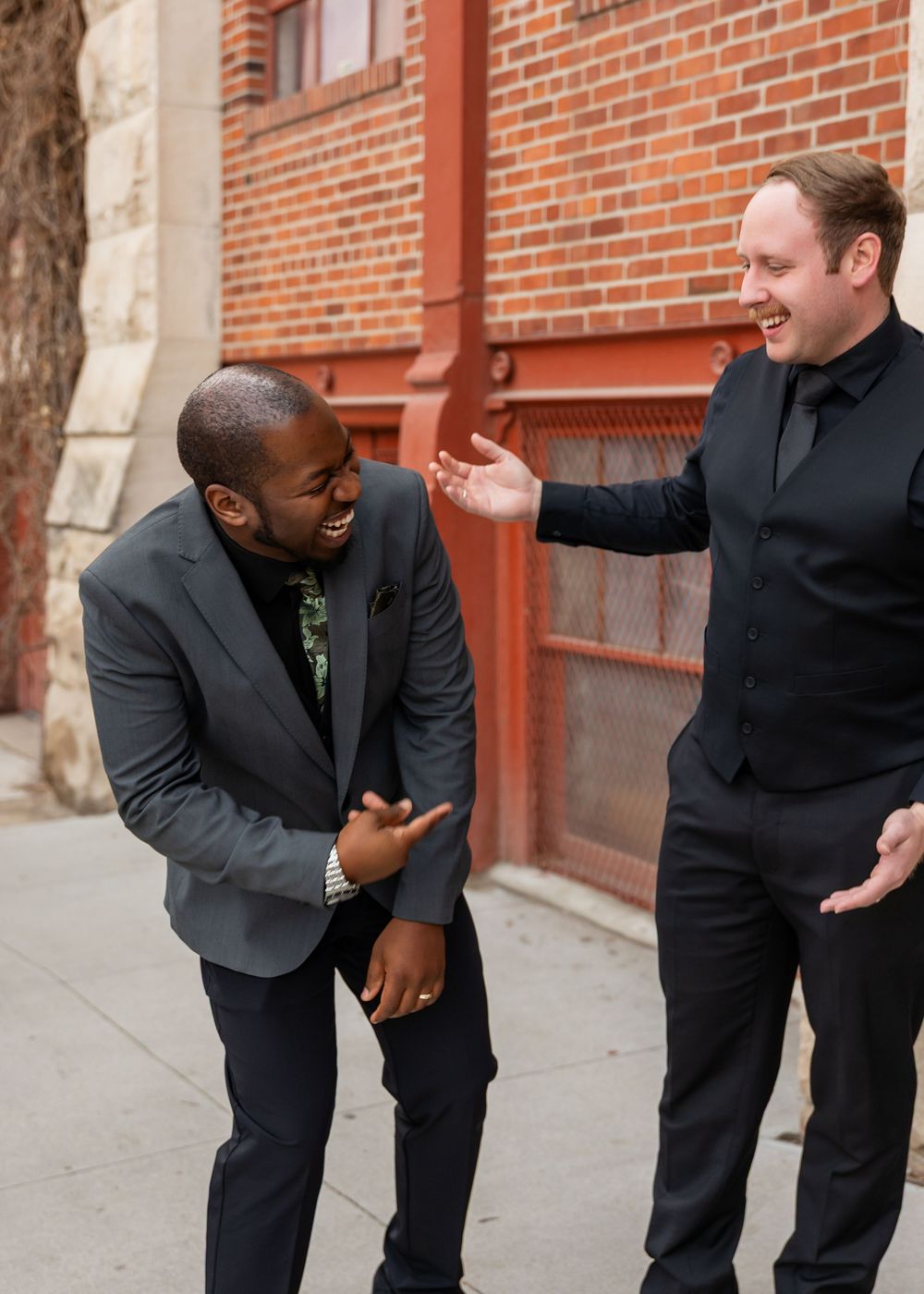 Two men in black suits laugh together outside a red brick building.