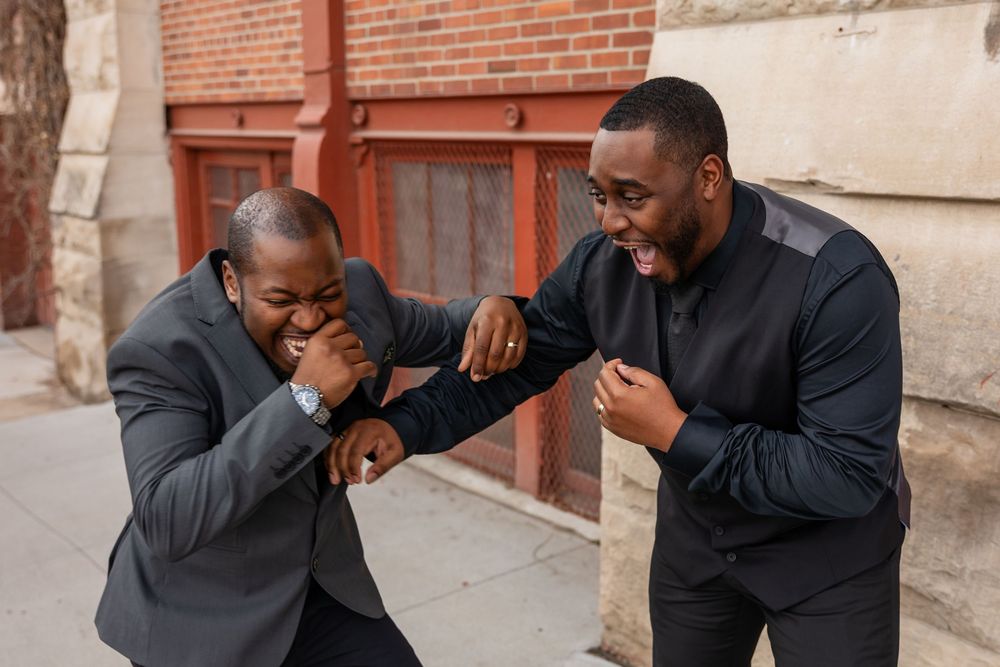Two men in suits playfully sparring and laughing on a city sidewalk near a brick building.