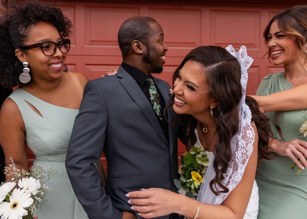Bride and groom share a joyful moment surrounded by smiling bridesmaids in sage green dresses at a wedding.