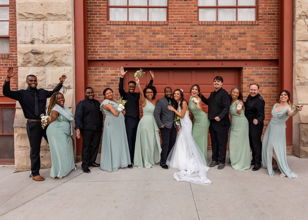 Wedding party poses outside brick building, bridesmaids in sage green gowns with groomsmen in black suits.